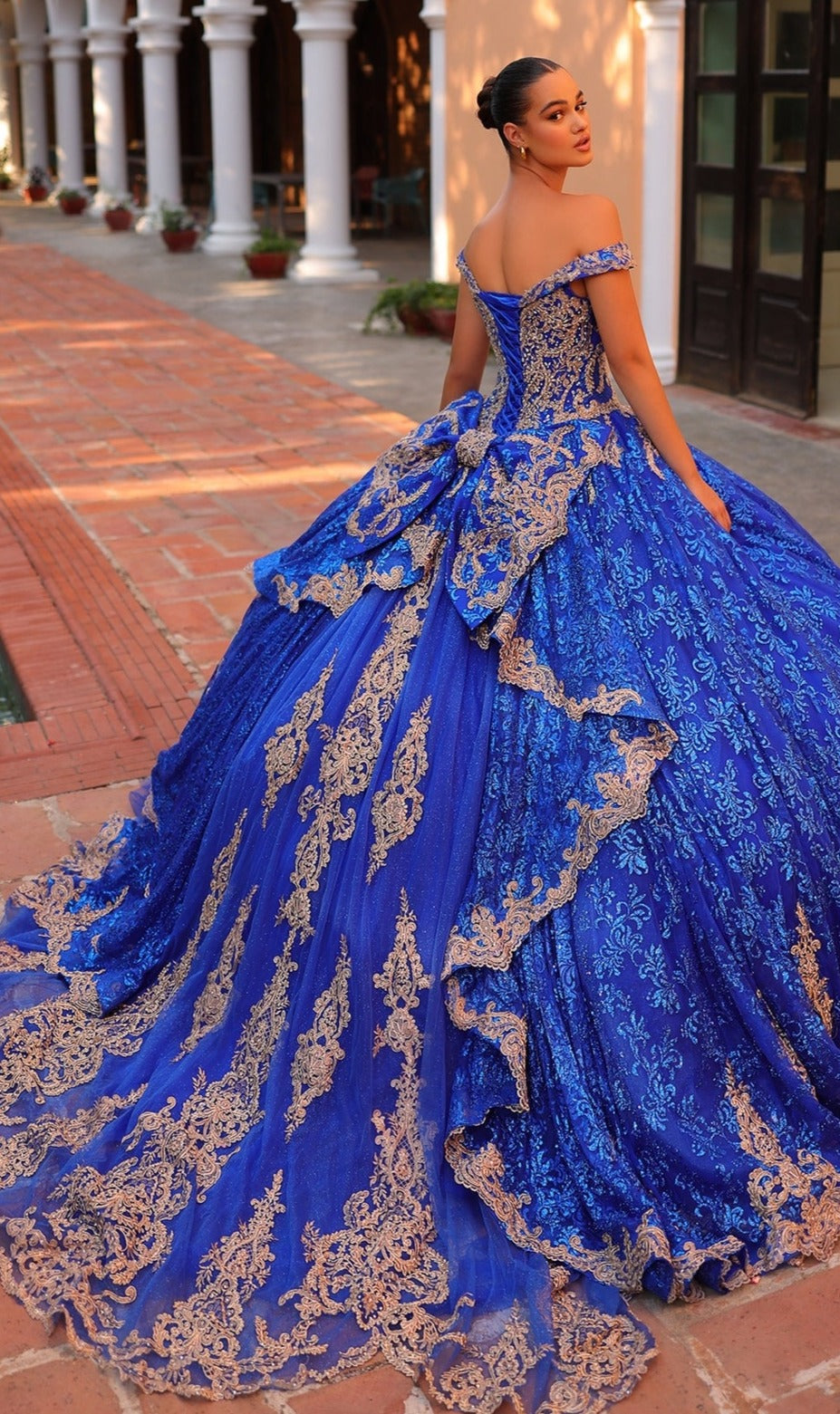 A woman stands outdoors in the Quinceanera Dress 54324 By Amarra, a stunning blue and gold ball gown featuring intricate lace embroidery, a long ruffled train, and an elegant off-the-shoulder design.